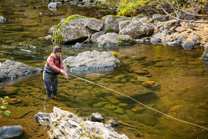 An angler in waders casts her fly line down a rocky river.