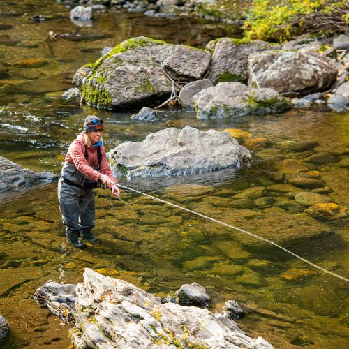 A woman in waders casts her fly rod along a rocky river.