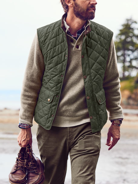 Man in Chocolate Pure Cashmere Simoom Sweater and Dark Green Barbour® Lowerdale Gilet walks along the beach.