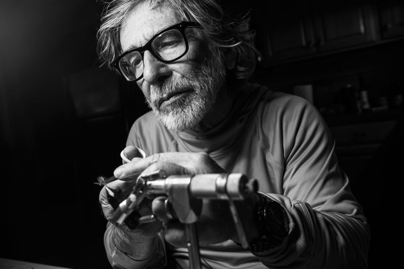 A black-and-white image of a silver-haired man tying a fly on a vise.