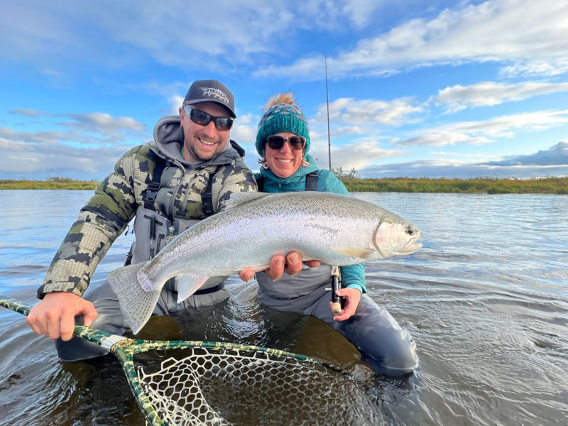 A man and a woman kneeling in a river and holding up a fish to the camera.