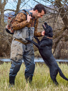 A man standing in a field wearing waders looking at a black lab