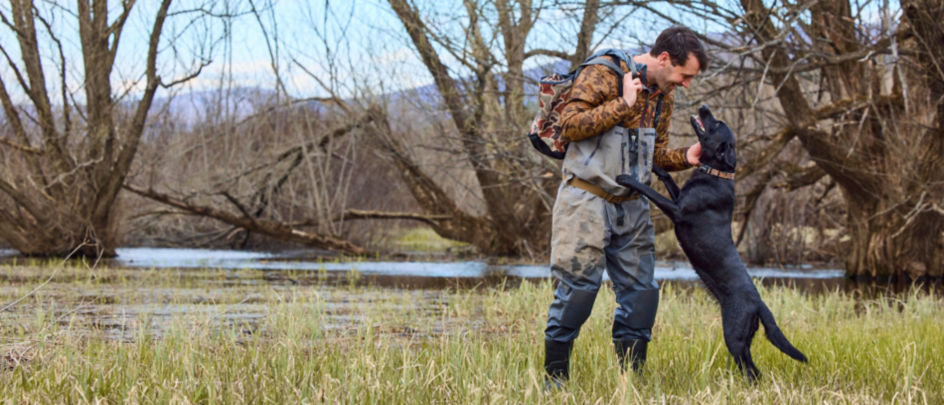 Charley Perkins wearing a camo-patterned jacket pets his black Labrador Retriever in a marsh