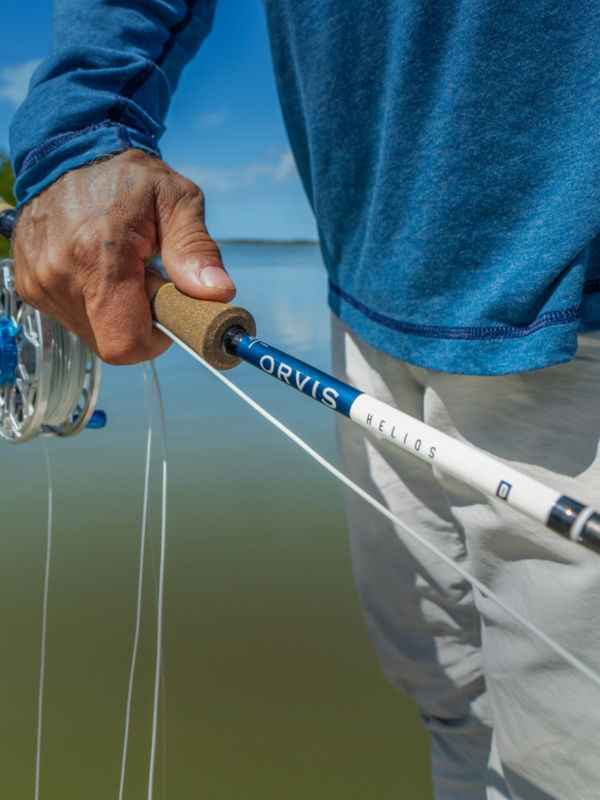 A hand gripping the butt of the Orvis x Captains for Clean Water Helios fly rod with trees and water in the background.
