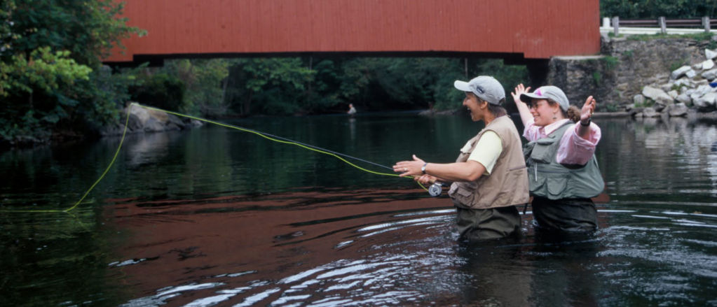 Two women fishing in a river in front of a red covered bridge