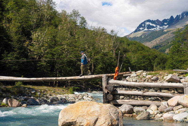 An angler carries a fly rod while crossing a rough bridge over a river.