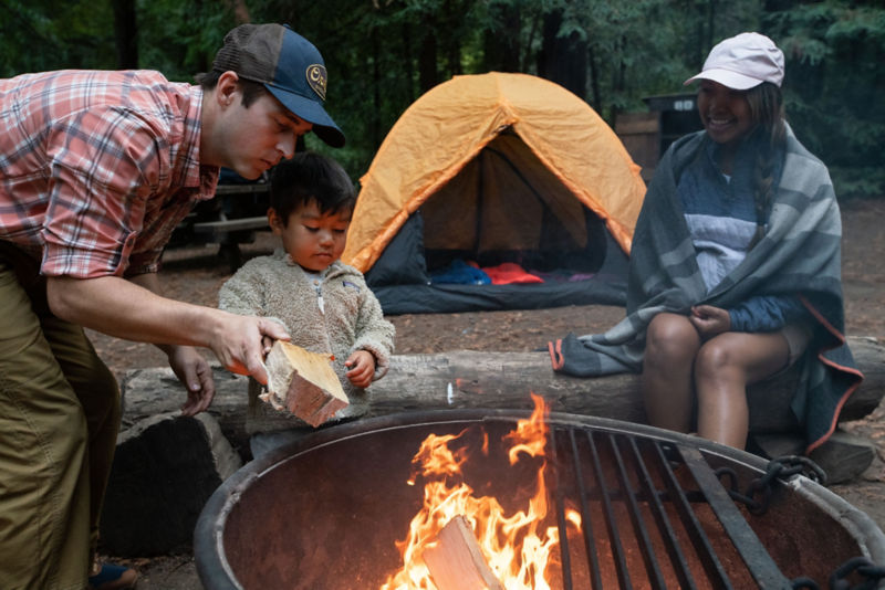 A father helps a child add wood to a fire while a mother watches.