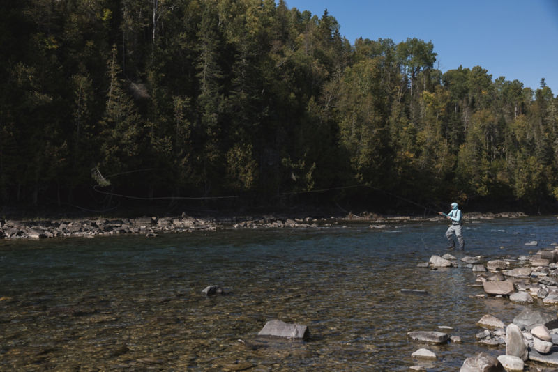 An angler stands on the side of a shallow rocky river and casts into a deep pool.