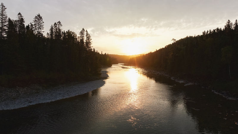 The sun setting over a river with pines on either side of rocky beaches.