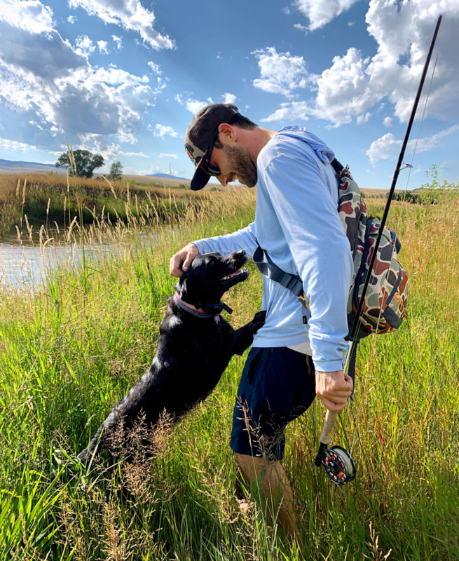 Charley pets Romi near the water while holding a fly rod.