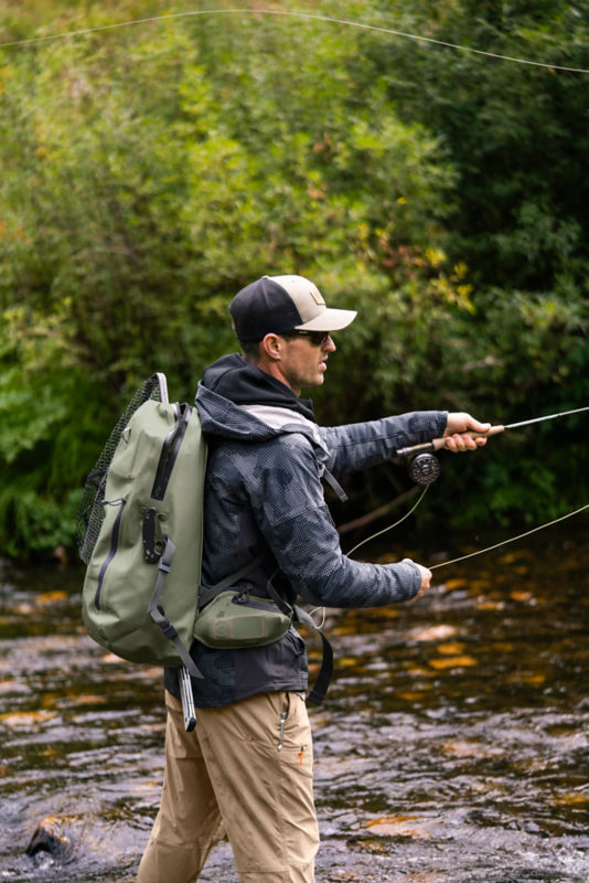 An angler wearing a PRO waterproof backpack throws line as he wades in a shallow, rocky river.