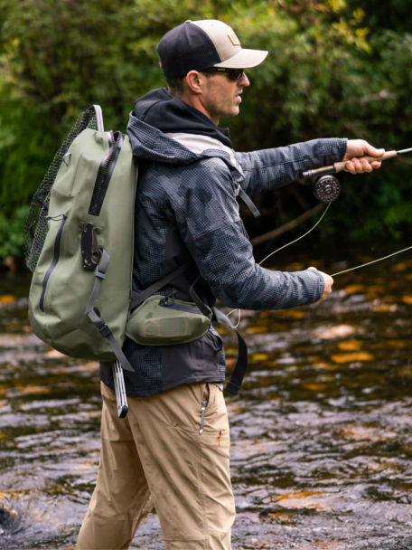 An angler wearing a PRO waterproof backpack throws line as he wades in a shallow, rocky river.