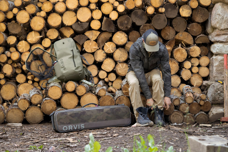 An angler puts on his wading shoes on a log pile with his pack gear off to the side.
