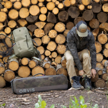 An angler puts on his wading shoes on a log pile with his pack gear off to the side.
