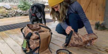 A woman wearing a yellow hat crouching down next to her black lab pouring food from a camp chuckwagon