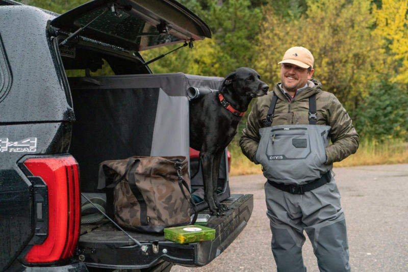 A man wearing waders standing by the back of his truck with his black dog peeking out of a gray travel crate.