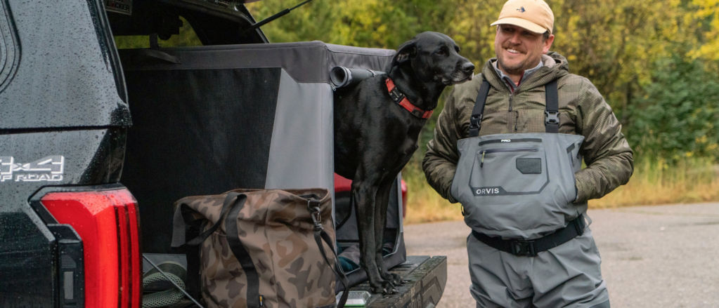 A man wearing waders standing by the back of his truck with his black dog peeking out of a gray travel crate.