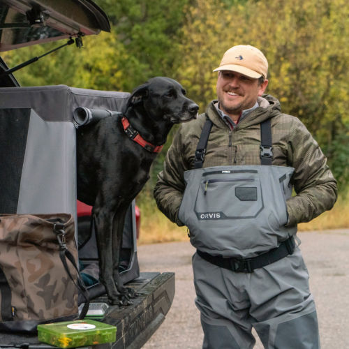 A man wearing waders standing by the back of his truck with his black dog peeking out of a gray travel crate