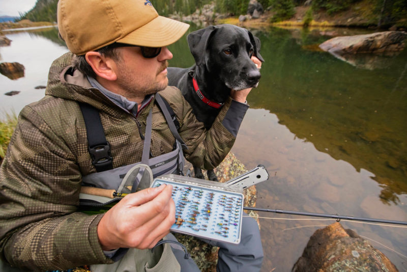 Man sits on river bank with his dog while wearing waders over his PRO Insulated Hoodie.