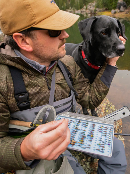 Man sits on river bank with his dog while wearing waders over his PRO Insulated Hoodie.