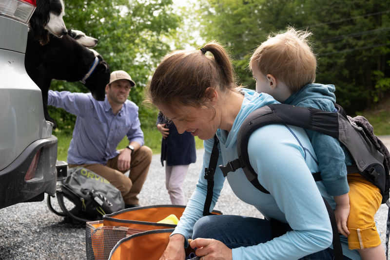 A mom with a child in a carrier on her back, sorts through a nylon tote bag.