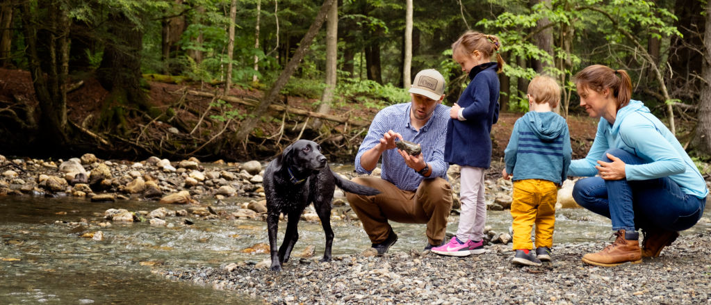 A family of two parents, two young children, and their black Labrador Retriever crouch by a stream inspecting rocks.