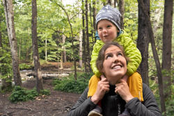 A woman on a walk with a child on her shoulders in the woods.