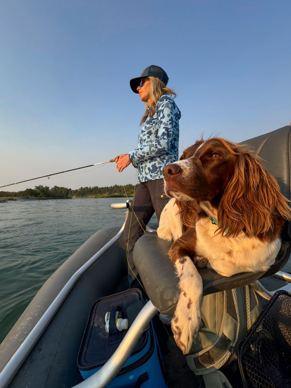 An angler in blue camo sun protection with her dog on a boat chair.