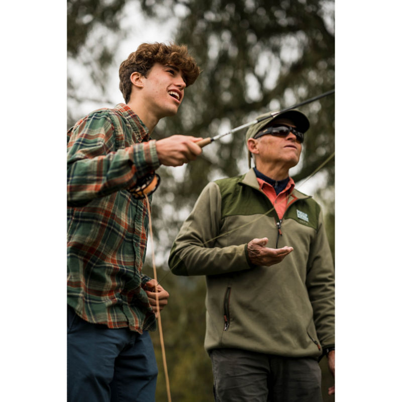 A young man learns how to cast; an older instructor behind him offers guidance.
