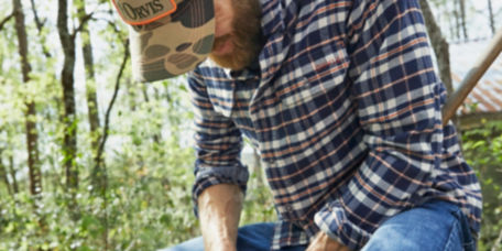 A bearded man in a flannel shirt works on a tool in the woods