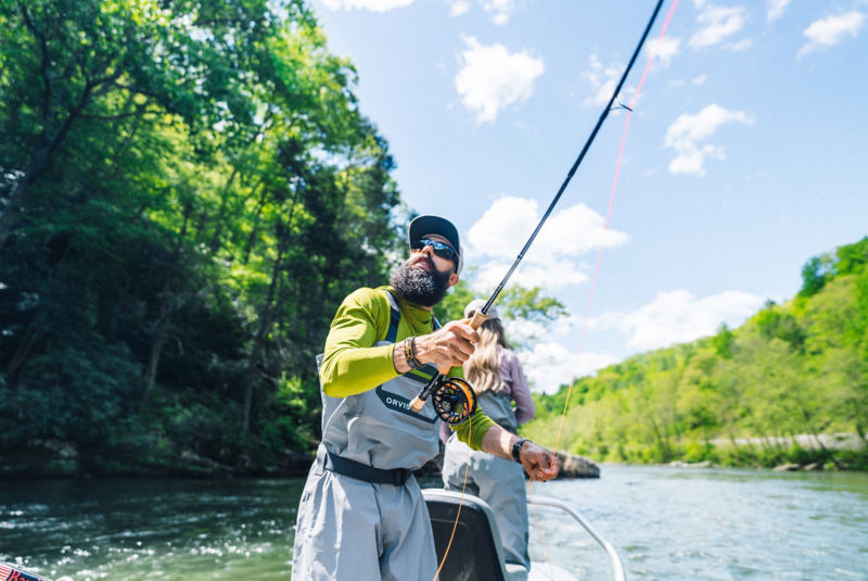 A sunglass-wearing angler casts from a small boat on a river under sunny skies.