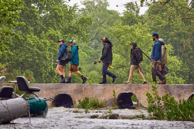 A group of anglers walk across a small bridge across a river with their equipment.