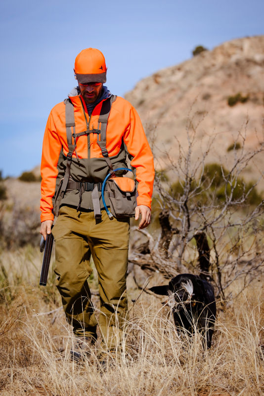 A hunter wearing a blaze orange jacket and tan pants with an upland vestholding a shotgun.