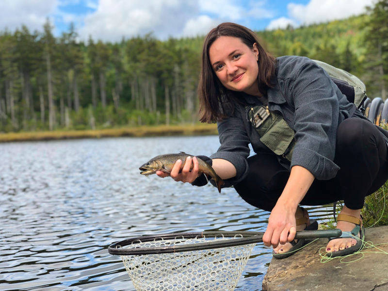Erin Heiny shows off a fish by the side of a forested lakeErin Heiny shows off a fish by the side of a forested lake