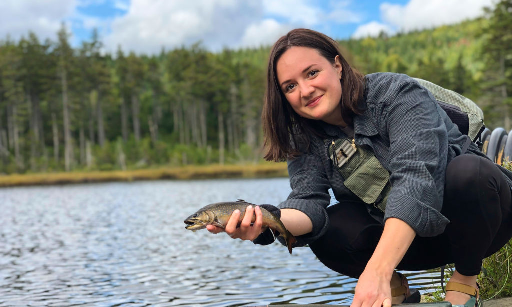 Erin Heiny shows off a fish by the side of a forested lake