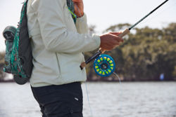 An angler wearing a brightly patterned fishing pack steadies their fly rod against their hip.