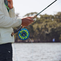 An angler wearing a brightly patterned fishing pack steadies their fly rod against their hip.