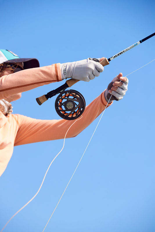 An angler in sun protection and Orvis gloves casts her line under a blue sky.