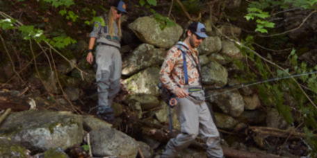 Two anglers in waders pick their way through rocks to a stream.