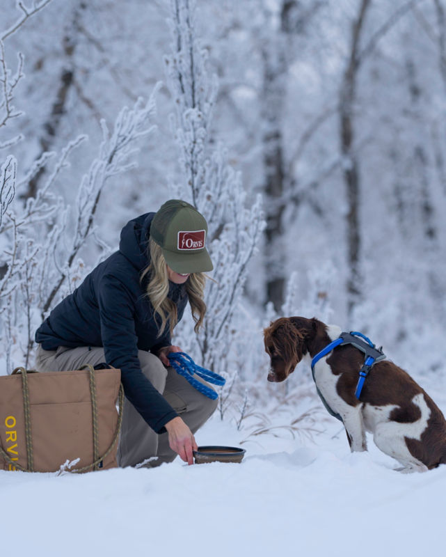 A smiling woman crouches in the snow to offer a travel bowl of food to her pup.