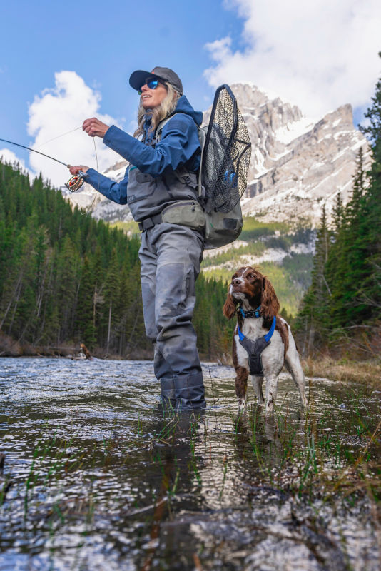 An angler casting from ankle-deep in a river accompanied by her spaniel
