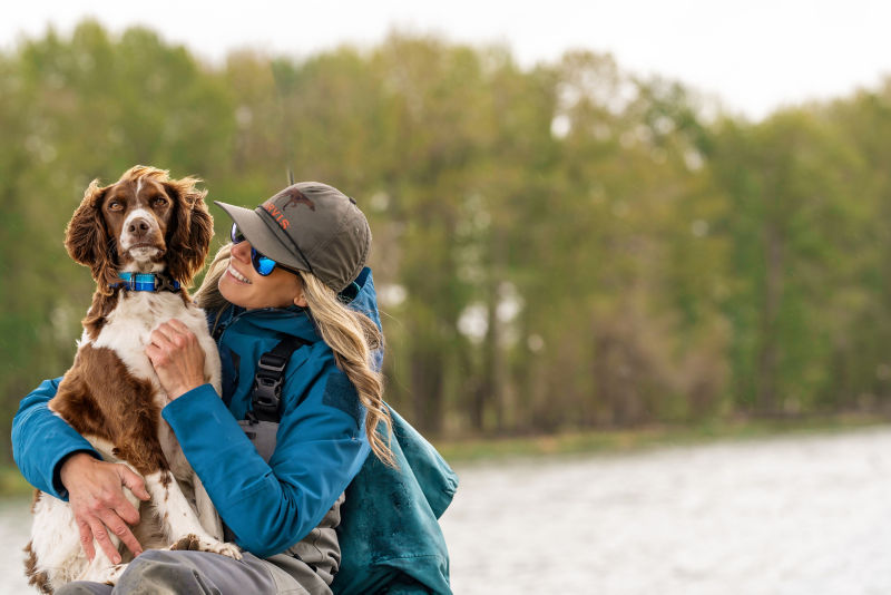 Woman holding her dog on the side of a river.