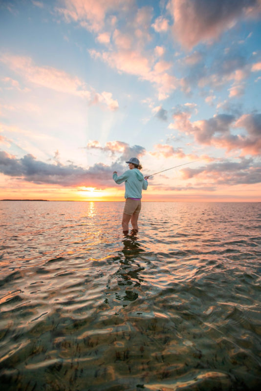 An angler casts from calf-deep in the ocean with a gorgeous sunrise behind her.