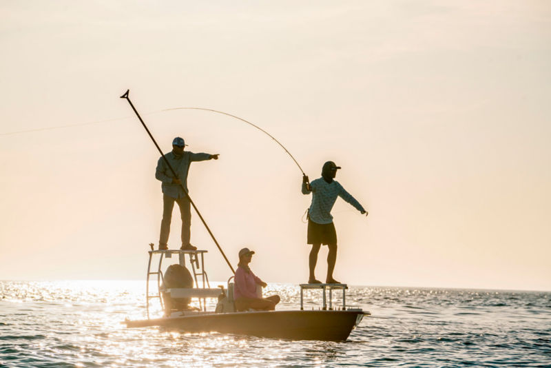 An angler in a boat casts while another poles a skiff through shallow waters at sunrise.