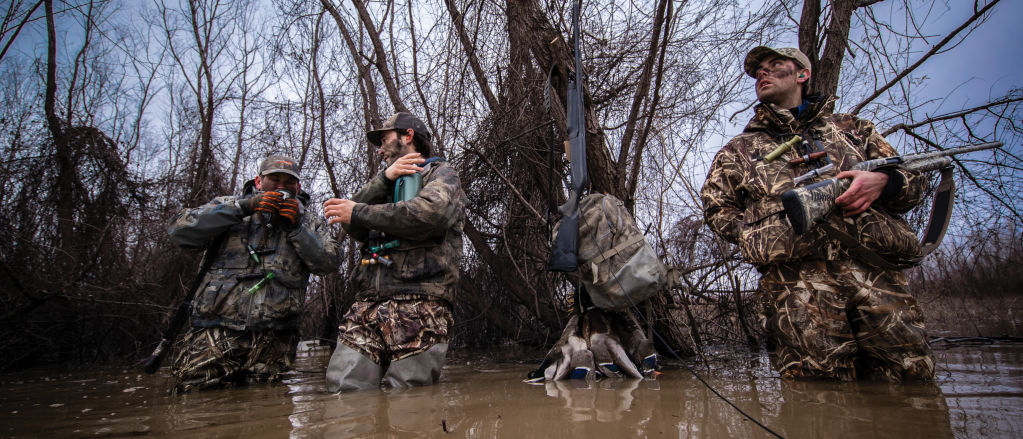 A hunter in camouflage stands waist-deep in water, holding his shotgun and looking towards the sky.