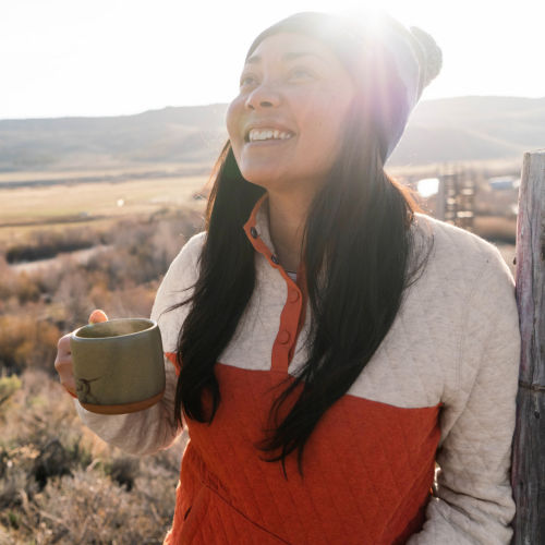 A smiling woman in a colorblock quilted sweatshirt holds a steaming mug