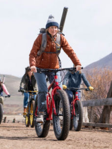 A woman riding a mountain bike with a Orvis pack on her back carrying a fly rod