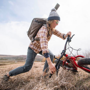 Woman wearing Bug-Out Backpack takes her bike off the ground against a cold countryside.