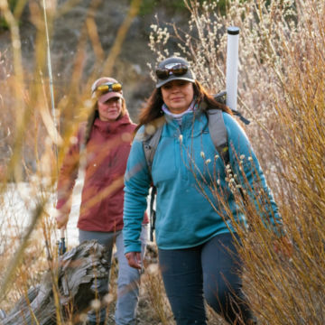 Two women in cold weather gear walk through high grass to a river.
