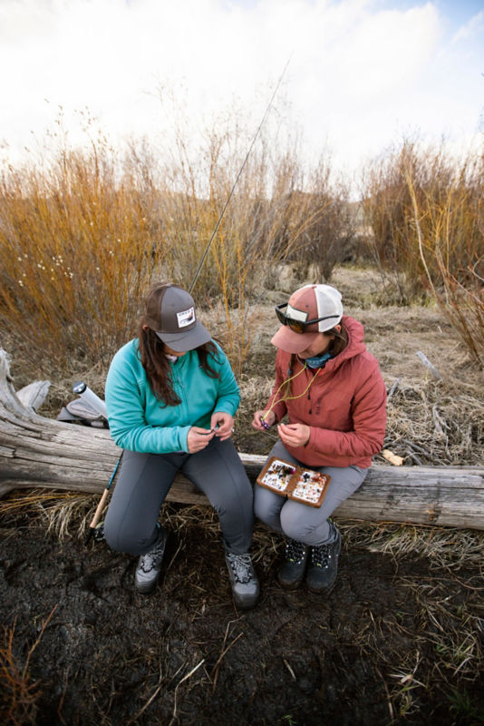 Two anglers sitting on a log choose which flies they'll use on the river